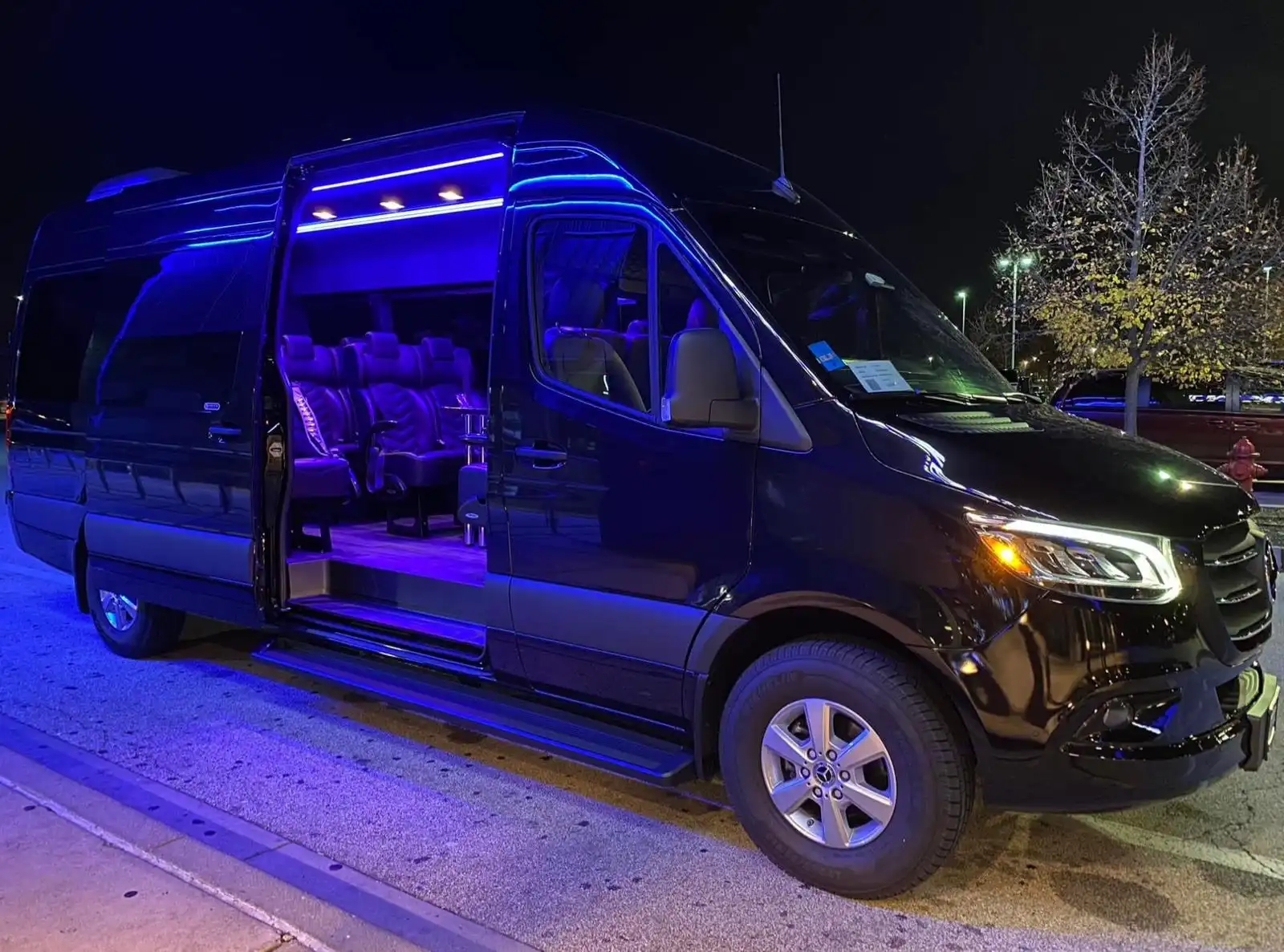 A black luxury Mercedes Sprinter van featuring spacious, high-end seating with blue ambient lighting.
